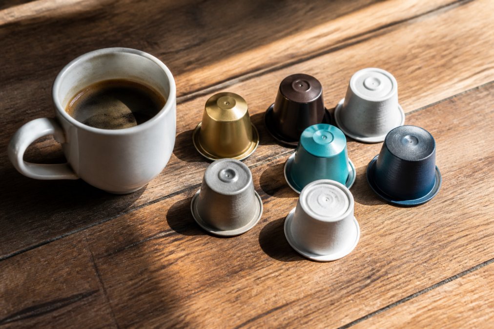 Flat-lay overhead shot of several coffee pods in different colors and materials arranged on a worn wooden surface next to a freshly brewed cup of coffee, with warm natural light casting soft shadows across the pod labels