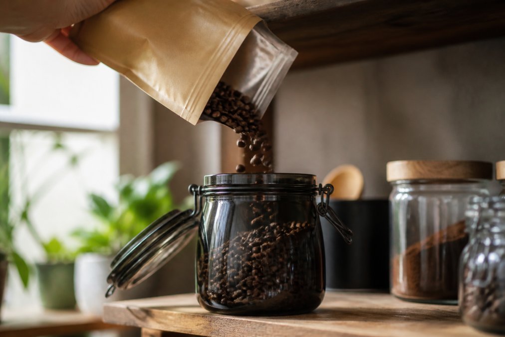 Close-up of whole coffee beans being poured from a kraft specialty bag into a dark, airtight glass canister on a wooden kitchen shelf, with soft natural light coming through a nearby window illuminating the beans