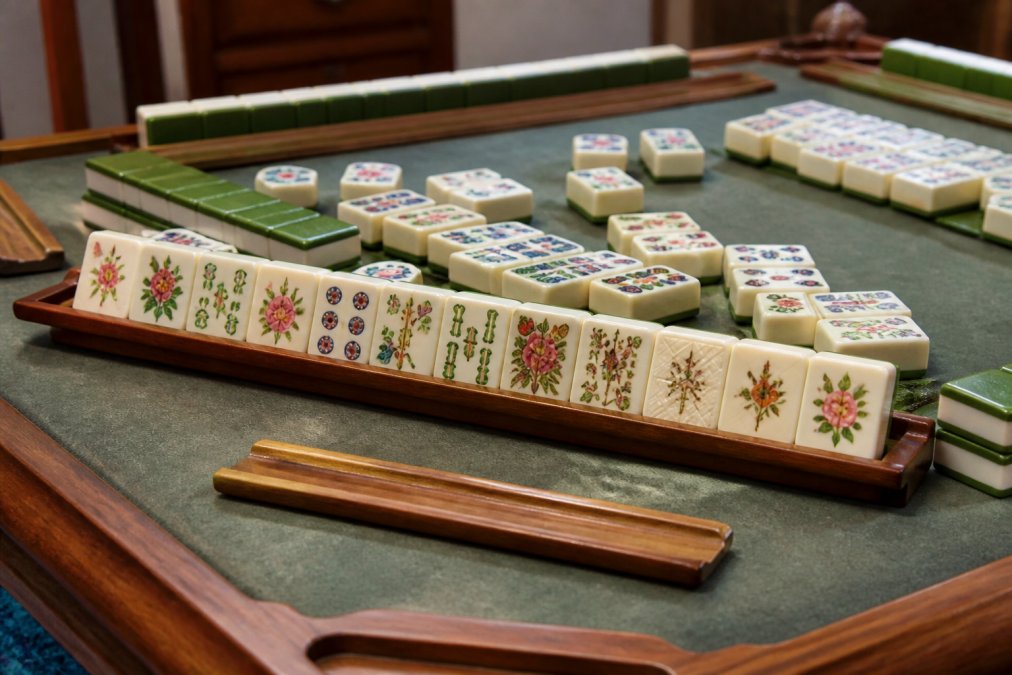 Close-up of colorful mahjong tiles arranged on a game table with wooden racks and pushers, showing vibrant hand-painted designs and intricate details under warm lighting