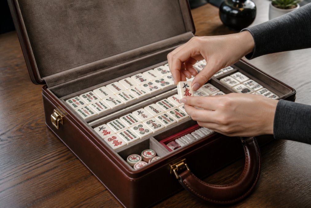 Hands carefully placing mahjong tiles into a premium leather carrying case with soft interior padding, demonstrating proper organization and protection of the complete set