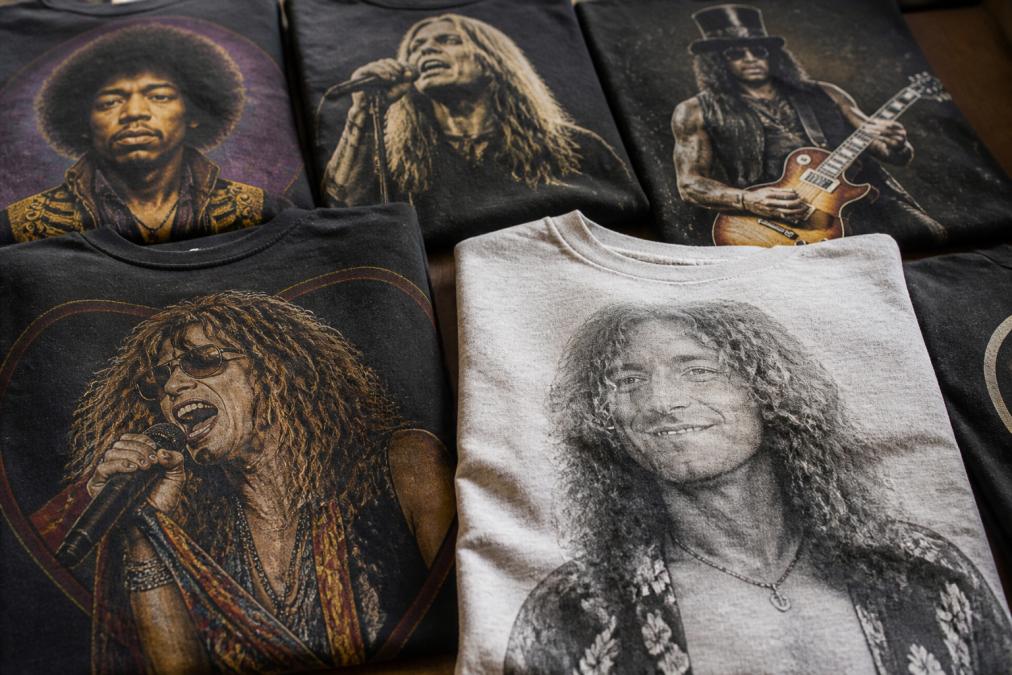 Folded collection of vintage-inspired band tees displaying original rock musician artwork with faded edges and warm-toned graphics on soft cotton fabric, arranged to show detail of screen printing and fabric texture