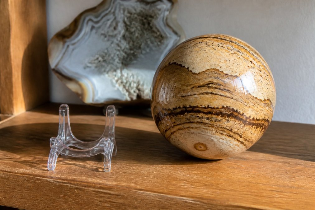 A polished picture jasper sphere resting on a wooden shelf beside a small acrylic display stand, warm natural light highlighting the landscape-like patterns in the stone