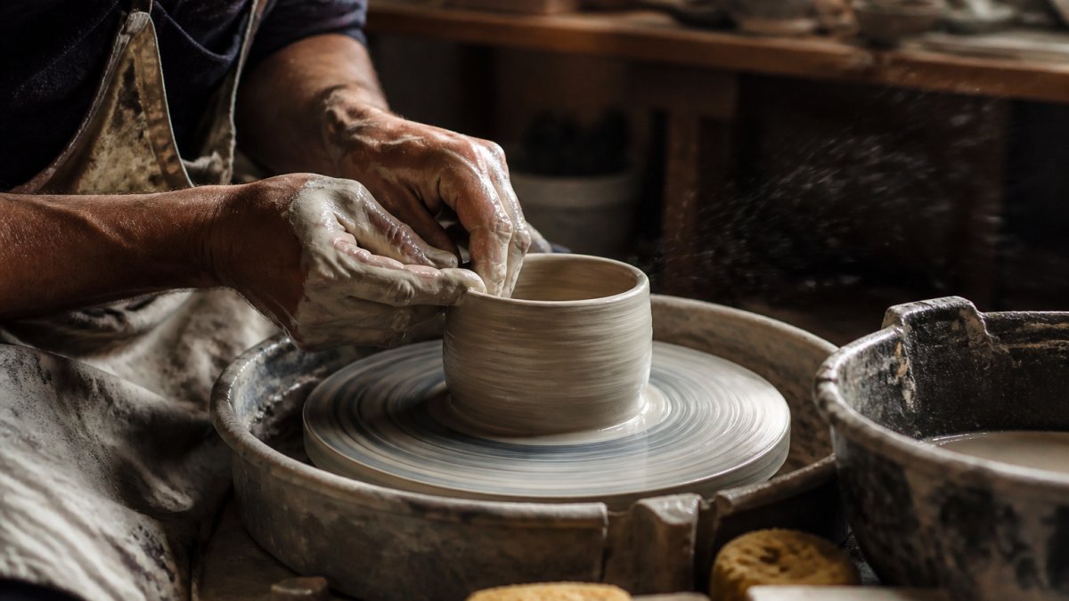 Close-up of a potter's hands shaping clay on a spinning wheel, fingers guiding the form with visible texture and water spray, natural studio lighting