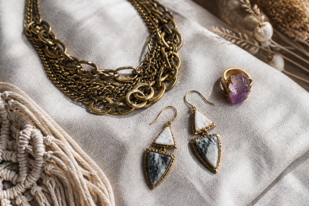 Flat lay of several handmade artisan jewelry pieces including a chunky layered chain necklace, raw gemstone ring, and bold statement earrings arranged on a linen cloth with soft natural lighting
