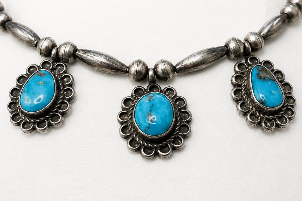 Close-up of a handcrafted squash blossom necklace displaying intricate sterling silver work with multiple blossom-shaped pendants and turquoise centerpiece against a neutral background, showing detailed metalwork and stone setting