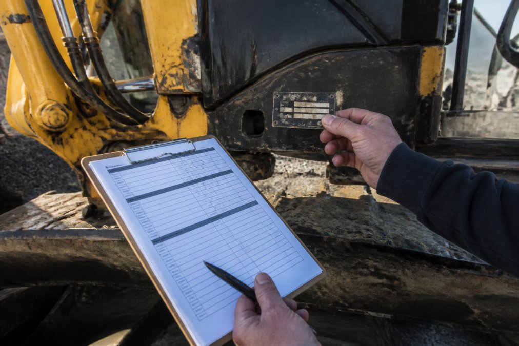 Hands holding equipment documentation and serial number verification form next to the excavator's nameplate in bright daylight, with clipboard and pen visible