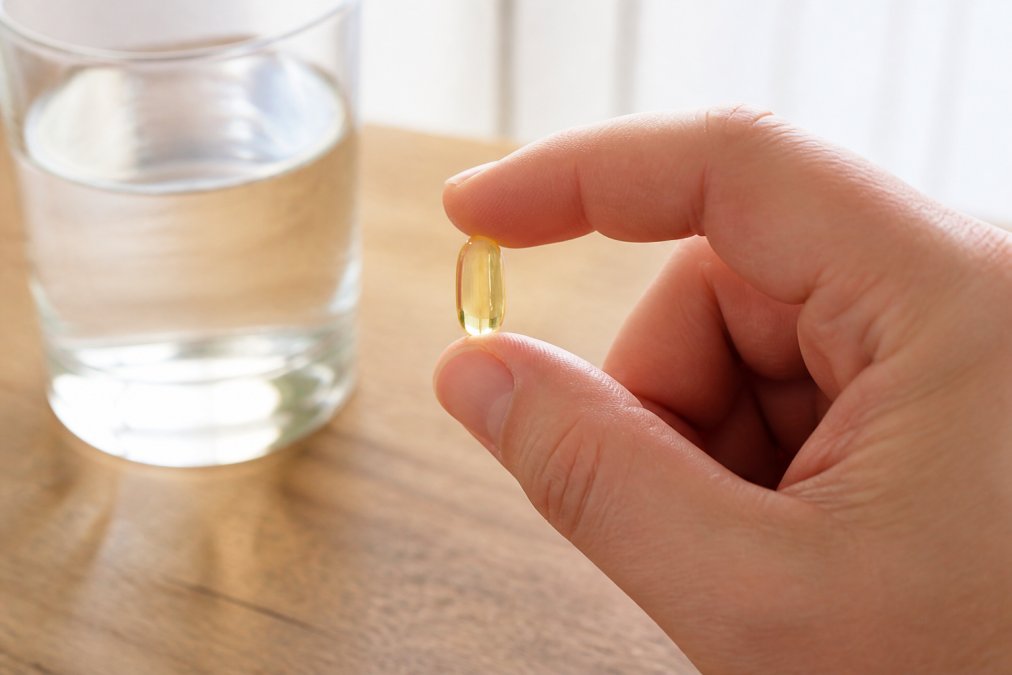 Close-up of a hand holding a single clear capsule supplement between thumb and forefinger against a bright white background, with a glass of water and a wooden surface visible in soft focus