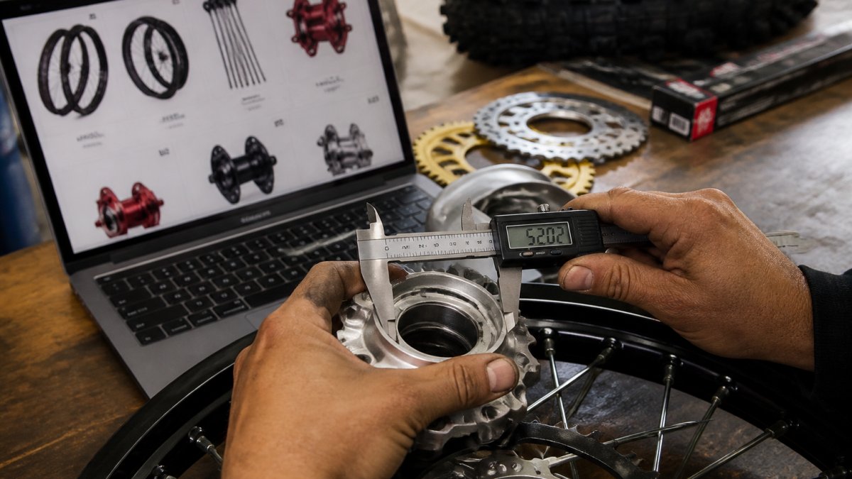 A close-up photo of a mechanic's hands holding a digital caliper against a metal dirt bike wheel hub, with a laptop showing an online parts catalog in the background
