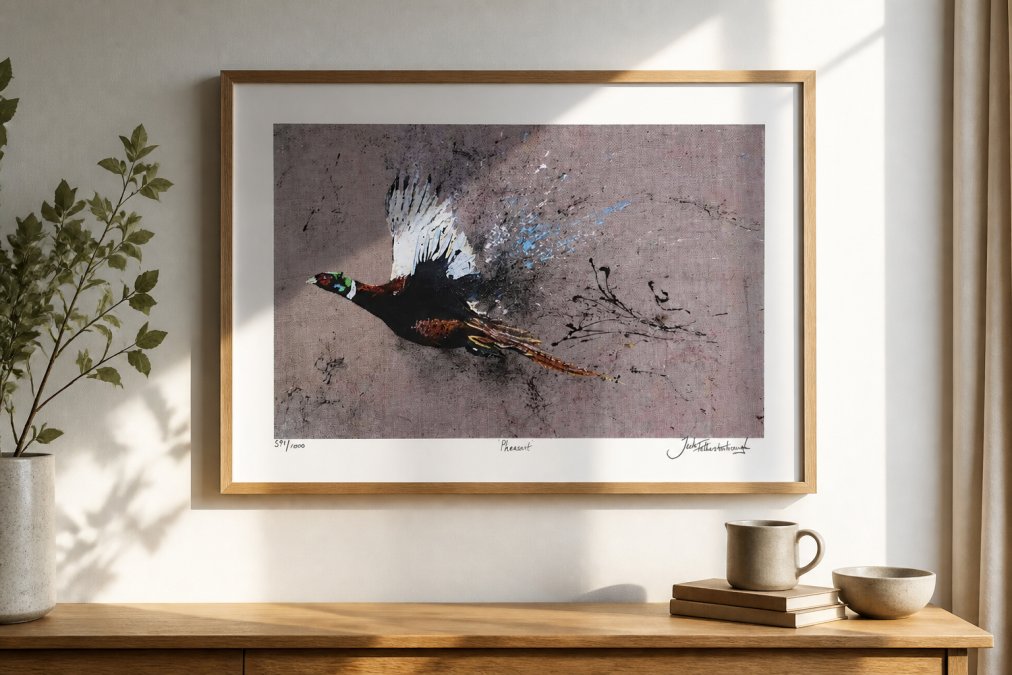Close-up of a framed pheasant wildlife art print displayed on a light-coloured wall above a wooden sideboard, with natural afternoon light highlighting the brushwork and pencil signature at the bottom of the print