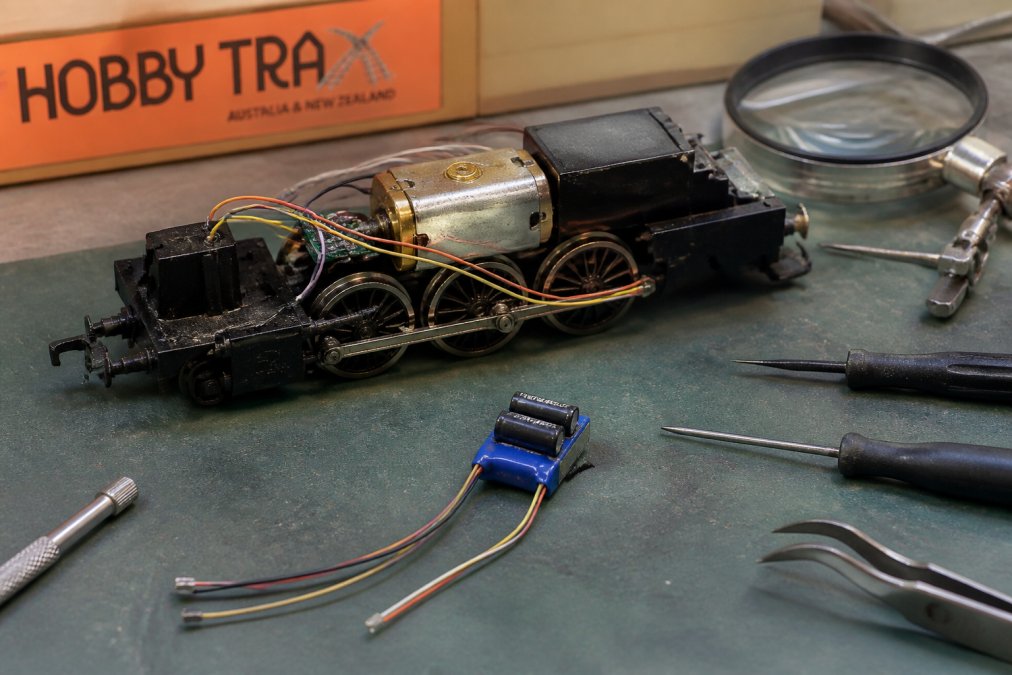 Model railway locomotive chassis on a workbench with a stay-alive capacitor module and decoder wires.