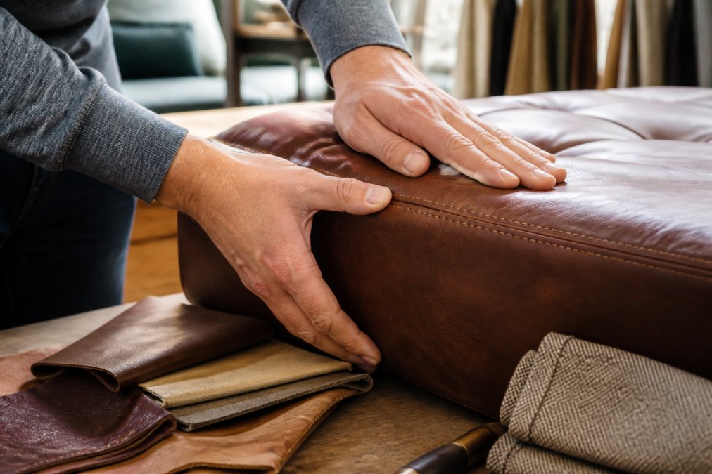 Close-up of hands examining aniline leather texture, running fingers along stitching detail and grain pattern on a luxury sofa sample in natural daylight