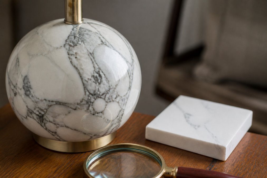 Close-up of a natural marble lamp base showing authentic veining patterns and texture, placed on a wooden surface next to a magnifying glass and a piece of faux marble sample for comparison