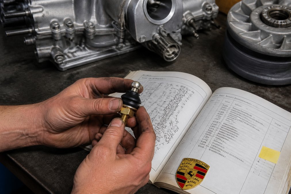 Technician's hands holding a vintage Porsche engine component next to technical documentation with part number labels visible under bright workshop lighting