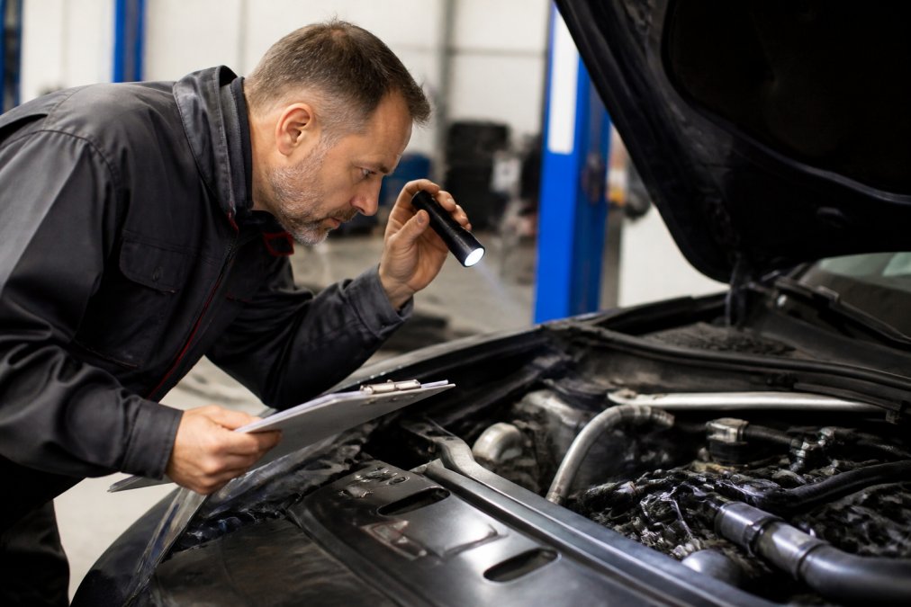 Professional mechanic examining used car's engine bay with flashlight and clipboard, hands checking under the hood with diagnostic tools visible