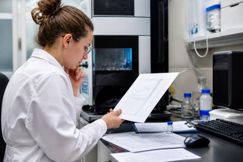 A laboratory technician examining analytical test results on a computer screen in a modern research facility, with printed batch reports visible on the desk