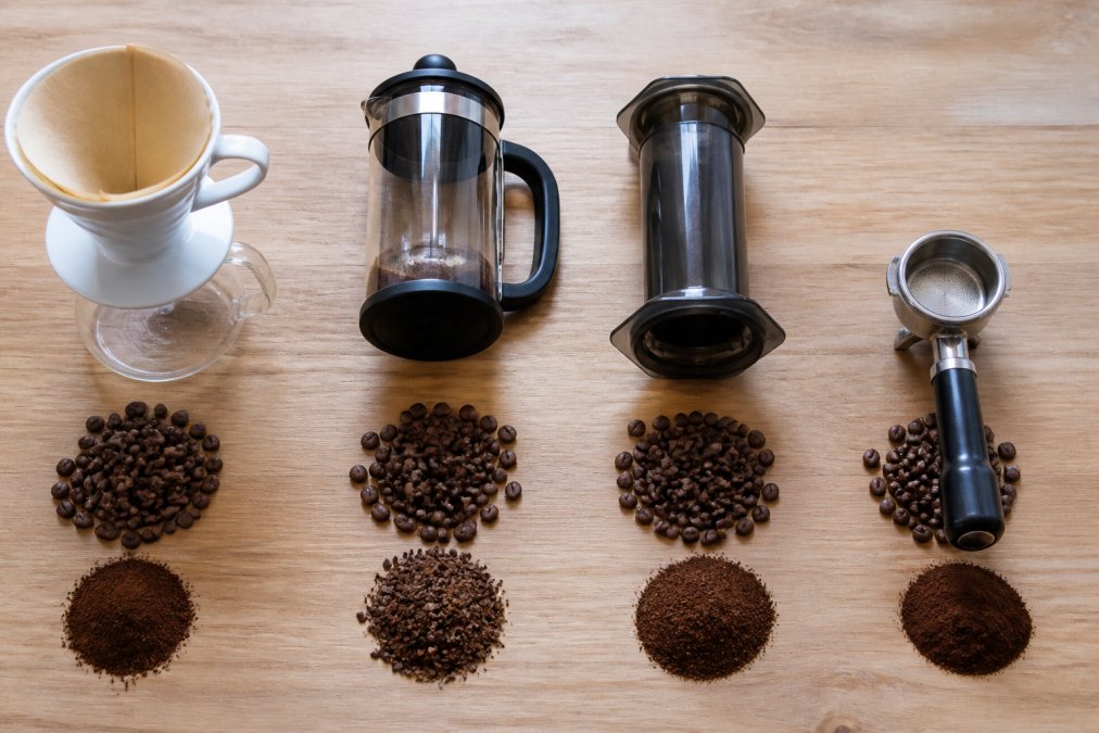 Flat lay of different brewing equipment arranged on a light wood surface: a pour-over cone with filters, a French press, an Aeropress, and an espresso machine portafilter, each surrounded by whole beans and a small pile of ground coffee at the appropriate grind size