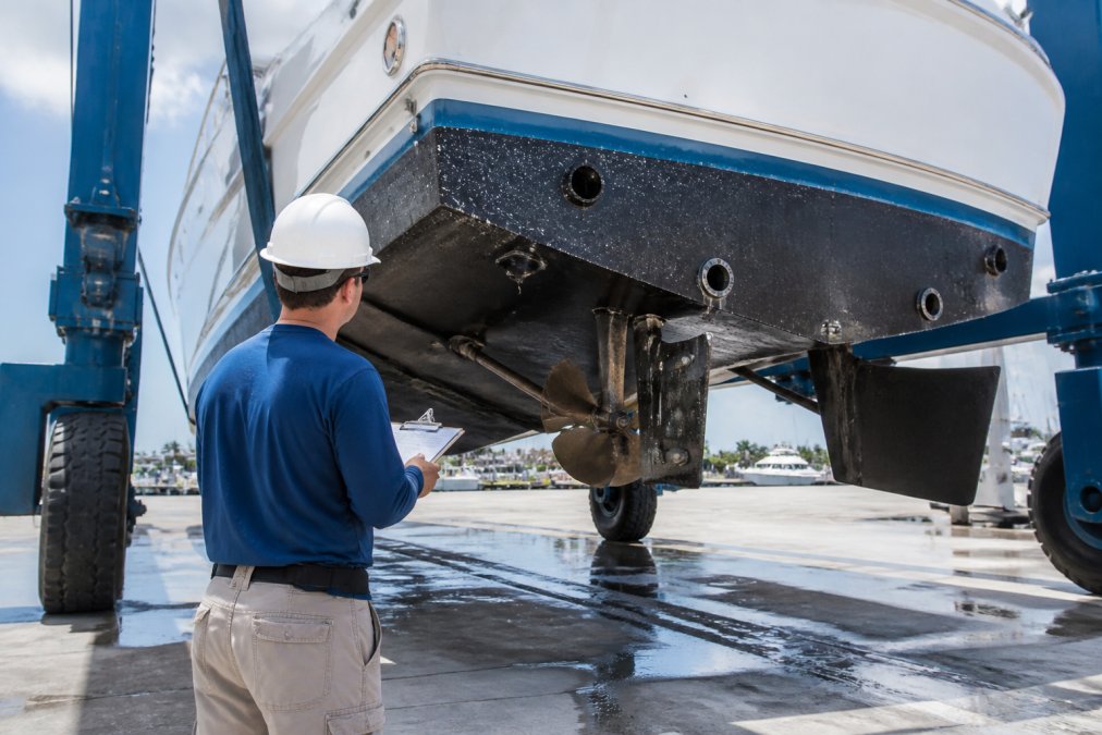 A marine surveyor in work clothes inspecting the hull and through-hull fittings of a large sportfish yacht hauled out on a travel lift at a boatyard, clipboard in hand, with the yacht's running gear visible