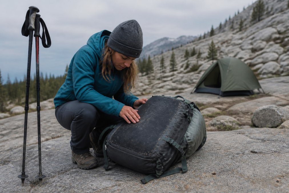 A hiker crouching on a rocky granite trail examining the reinforced abrasion panel on the bottom of a loaded backpack, trekking poles planted beside them and a pitched tent visible in the background under overcast light