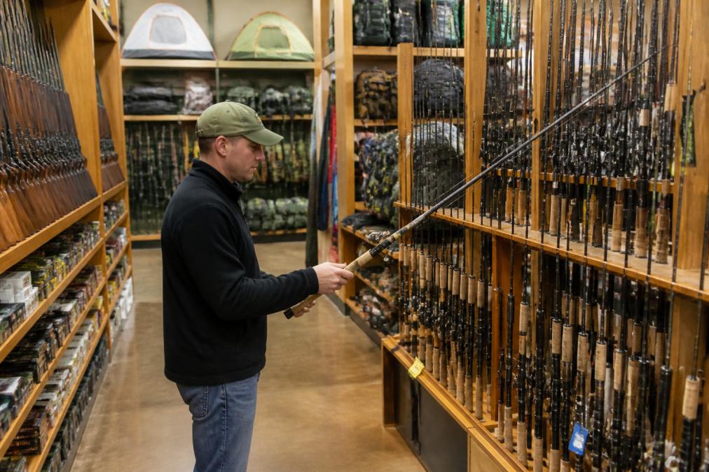 Outdoor enthusiast examining fishing rods, hunting rifles, and camping equipment on retail shelves with multiple brand options visible, natural store lighting