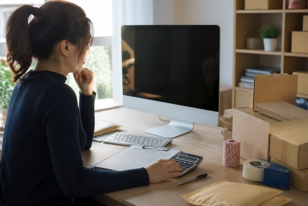 E-commerce business owner sitting at desk reviewing multiple freight quotes on a computer screen, with a calculator, clipboard with shipment details, and packaging samples visible on the desk, bright office lighting