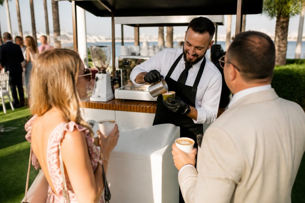 Professional barista crafting specialty espresso drinks for event guests at outdoor venue, with mobile coffee cart in background and attendees holding custom beverages