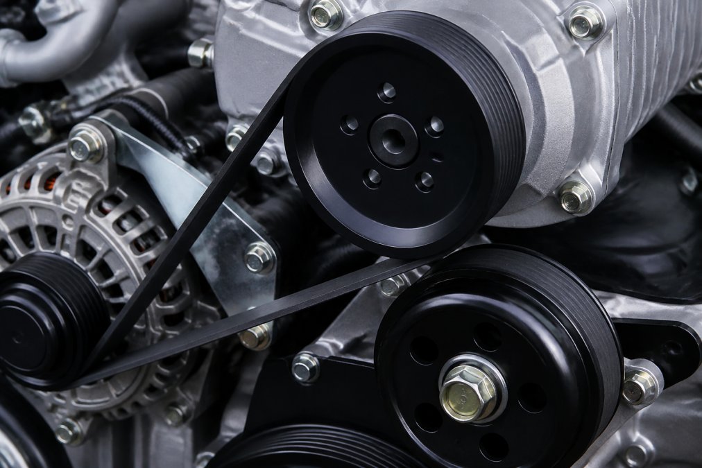 Close-up of supercharger pulley system and belt drive assembly mounted on engine block, showing mechanical components and power transfer mechanisms in detailed workshop lighting