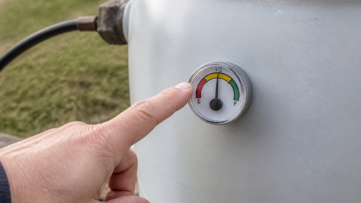 Close-up of a hand pointing to a propane tank pressure gauge mounted on the side of a metal cylinder, showing the fill level indicator needle positioned at approximately 50% capacity