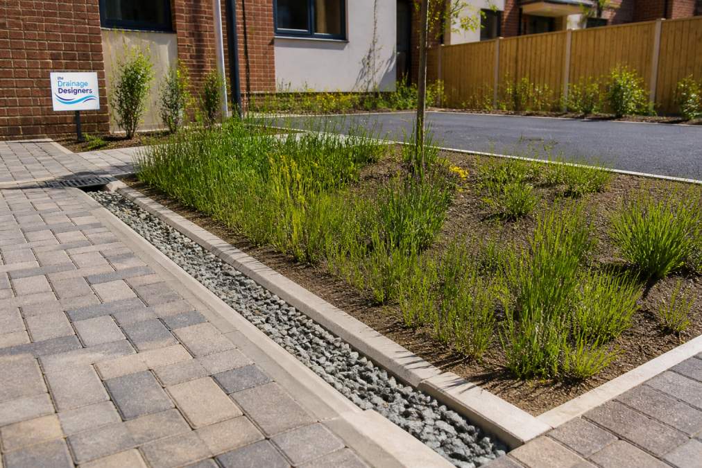 Sustainable drainage system installed on a small residential site showing permeable paving blocks with visible gravel sub-base, shallow drainage channel, and planted rain garden basin with native vegetation in afternoon sunlight