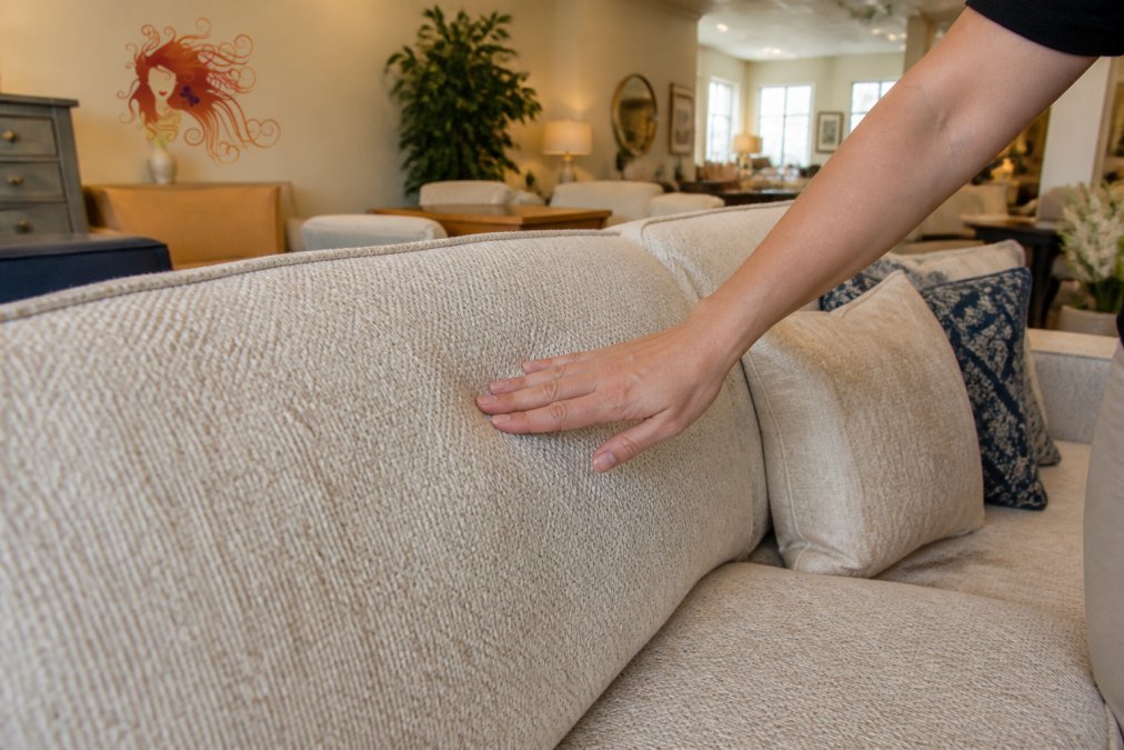 A close-up of a person's hand running over the fabric of a used sofa, highlighting the texture and condition in a well-lit consignment showroom