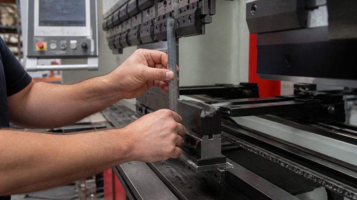 Close-up of a technician's hands holding a precision ruler against a press brake backgauge finger inside a metal fabrication shop, with the CNC controller screen visible in the background