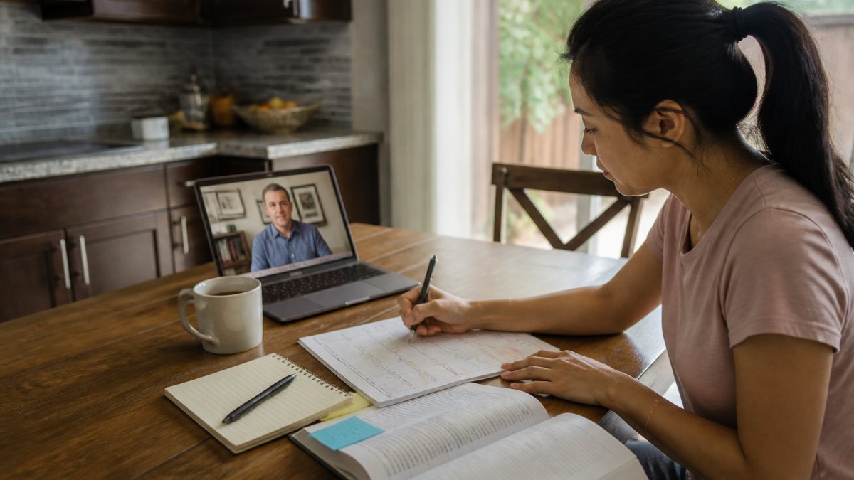 A person studying at a kitchen table with a printed calendar, a laptop showing an online course, a notepad, and a coffee mug, warm evening light