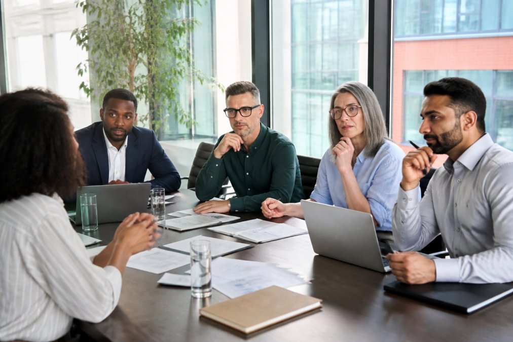 Diverse team of business professionals in a modern conference room during a pitch meeting, with laptops and presentation materials visible, discussing investment opportunities with natural office lighting