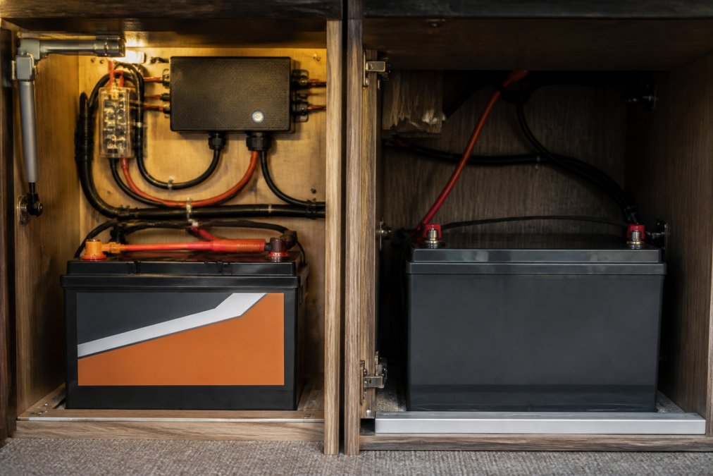 Close-up of a LiFePO4 lithium leisure battery installed inside a campervan electrical cabinet, with neatly routed wiring and a battery management system unit visible on a timber-clad panel under warm LED lighting