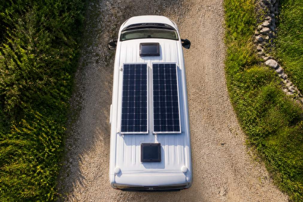 Overhead view of a white campervan roof with two rigid solar panels mounted neatly in parallel, parked on a gravel lane surrounded by green UK countryside under bright afternoon sunlight
