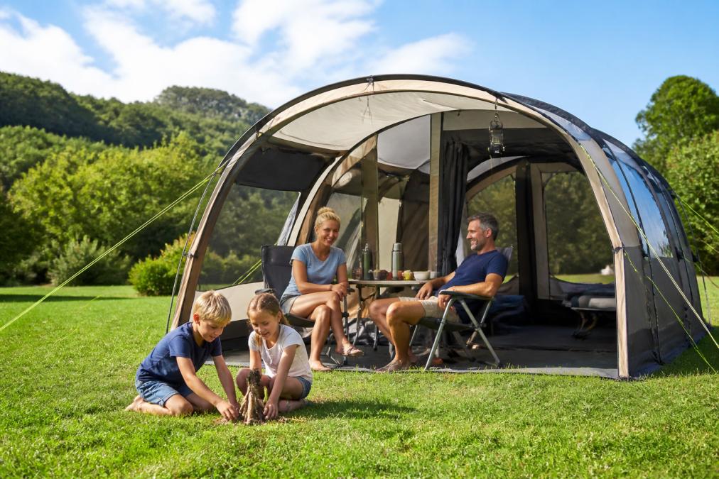 Vierköpfige Familie sitzt vor einem großen Familienzelt auf grünem Campingplatz, Kinder spielen davor, grüne Landschaft im Hintergrund, sonniger Sommertag
