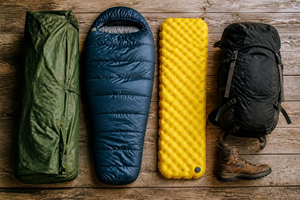 Overhead flat lay of a tent, sleeping bag, sleeping pad, and backpack arranged on a wooden surface with a hiking boot nearby