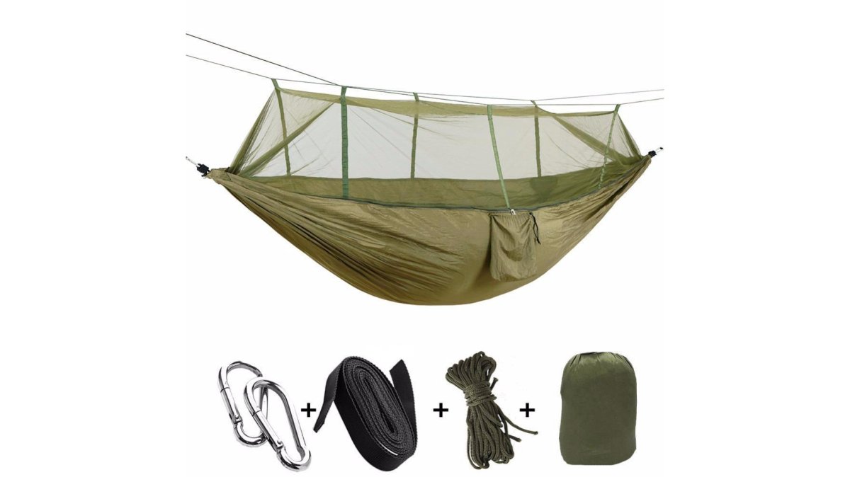 A flat lay of lightweight camping gear on a wooden table: a rolled navy sleeping bag beside a folded hammock with attached mosquito net and a small grey solar charger, with a sunlit bushland backdrop