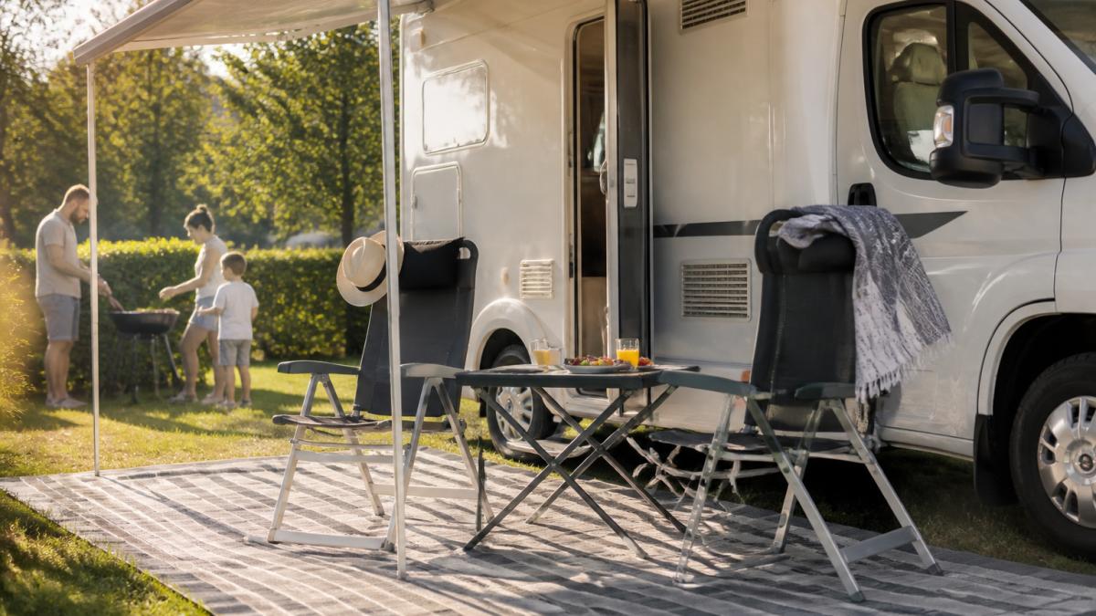 Ein Wohnmobil auf einem sonnigen Campingplatz, davor ein aufgebauter Vorzeltteppich und Campingstühle, im Hintergrund eine Familie beim Grillen