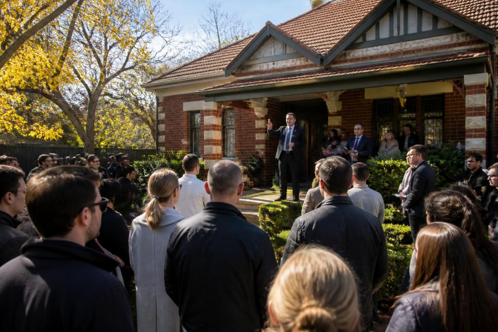 An outdoor Melbourne property auction scene with a crowd of bidders and an auctioneer standing in front of a period-style brick home on a sunny autumn day, natural daylight