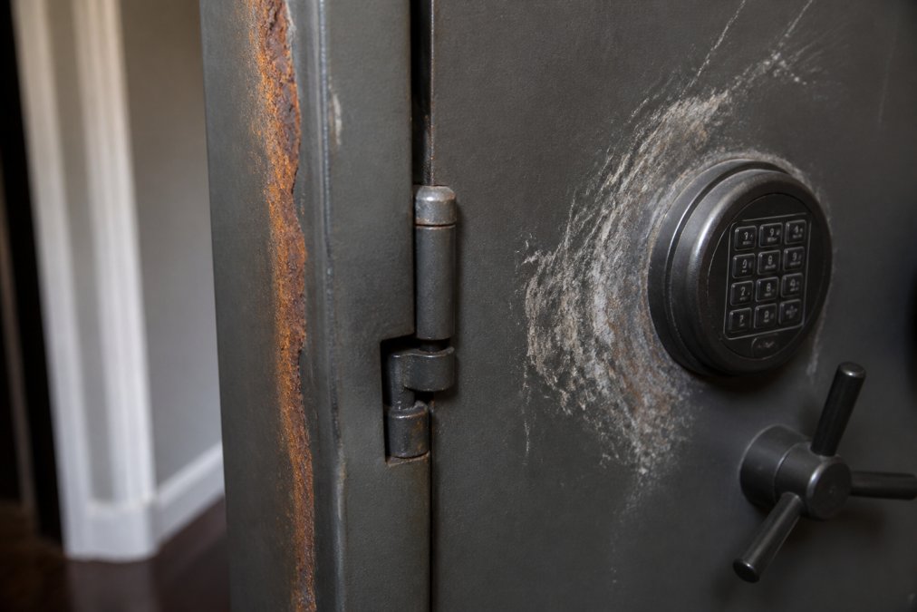 Close-up of a residential safe door showing visible rust along the edge, a slightly misaligned hinge, and scratches around the keypad from a forced entry attempt