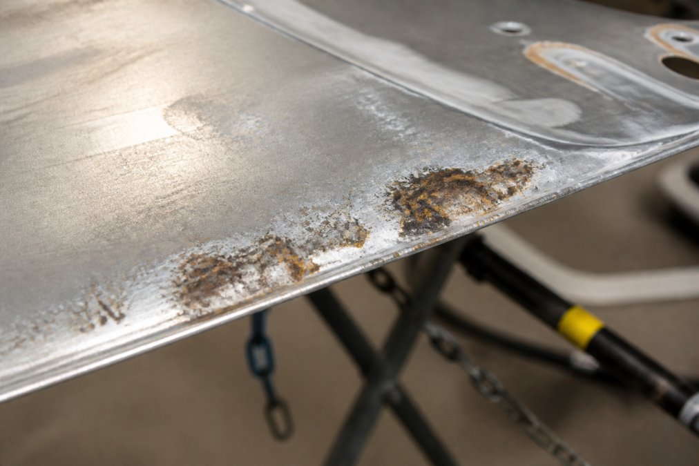 Close-up of corroded aluminum panel edge showing rust and oxidation damage from steel tool contact, with visible galvanic corrosion marks on the surface under workshop lighting