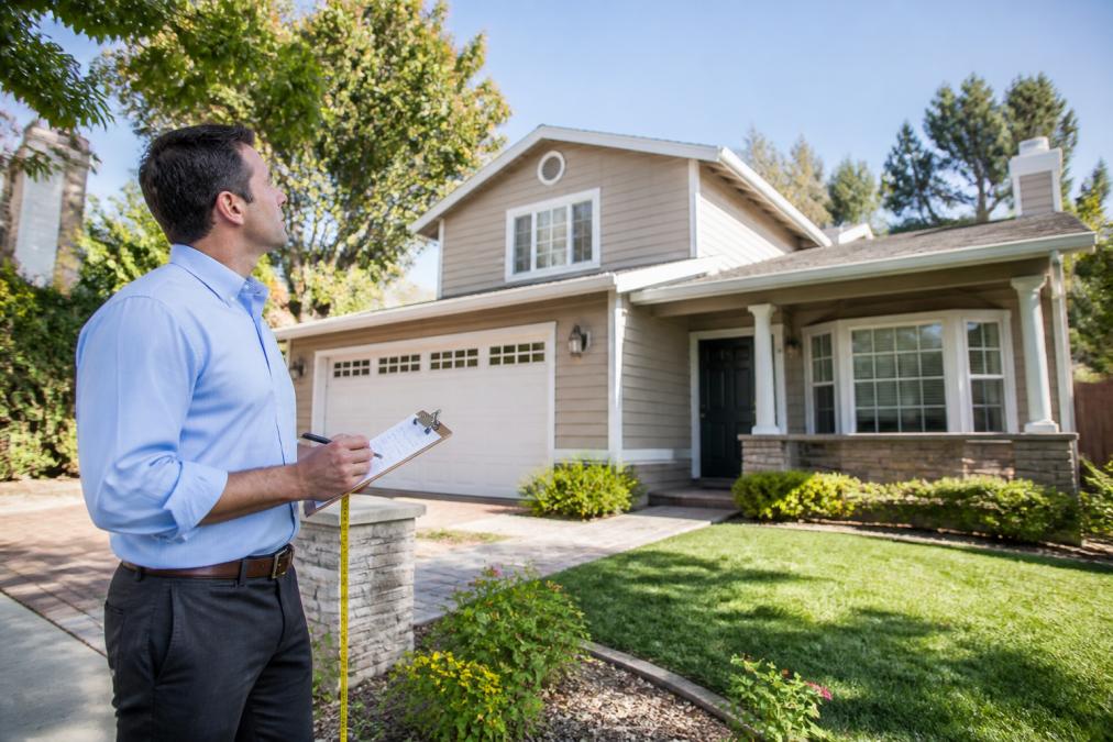 A professional appraiser in business casual attire inspecting the exterior of a well-maintained suburban house, holding a clipboard and measuring tape, with mature trees and a clear sky in the background