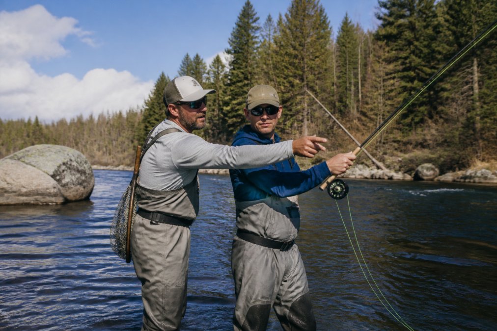 A fly fishing guide standing beside a client on a river, pointing at the client's casting arm and demonstrating a casting stroke with a fly rod in hand, with a scenic river and trees in the background