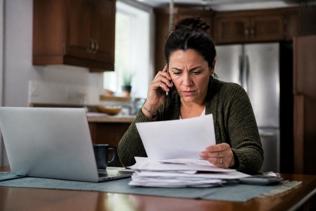A worried homeowner sitting at a kitchen table with a stack of mortgage statements, a laptop open, and a phone in hand, looking determined while reviewing options to stop foreclosure
