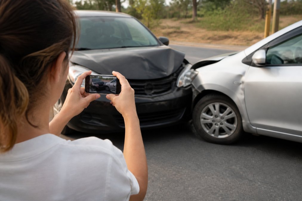 Person taking photos of vehicle damage at accident scene with smartphone, with damaged vehicles and road visible in background