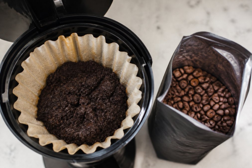 Dark espresso grounds inside the filter basket of a regular coffee maker next to a bag of whole coffee beans.