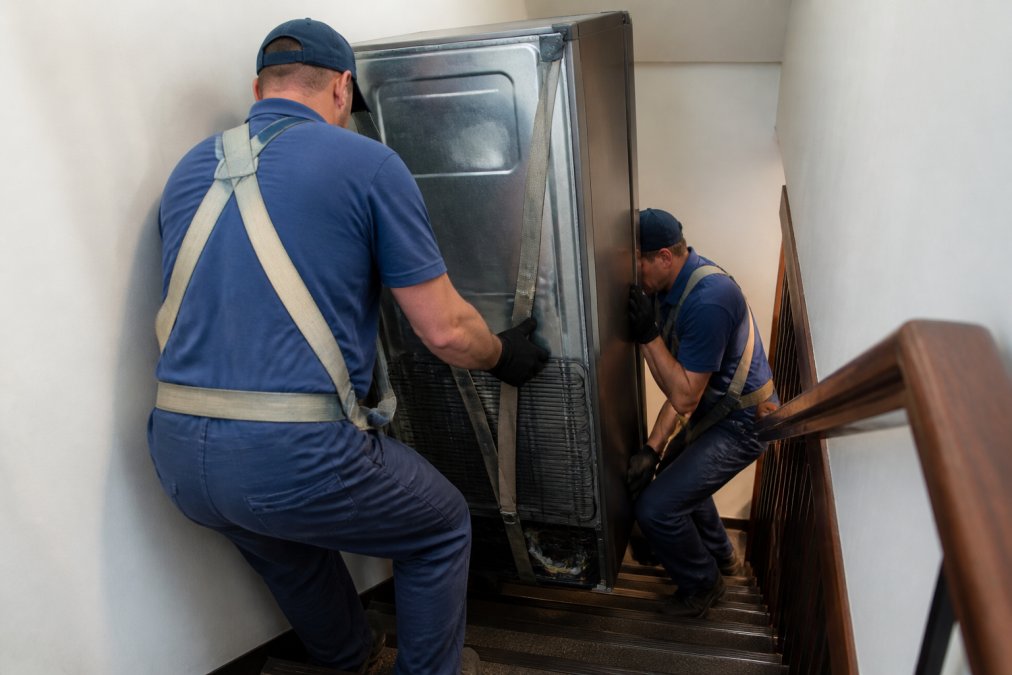 Professional two-person crew carefully maneuvering a large refrigerator down a narrow stairwell using proper safety equipment, lifting straps around the appliance, and coordinated body positioning with bent knees