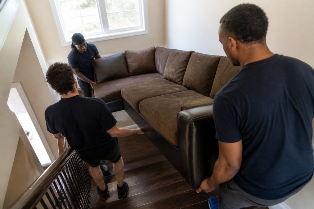 Professional junk removal team carefully maneuvering a large upholstered sectional sofa down a residential staircase, demonstrating coordinated lifting technique and proper hand placement, natural light streaming from stairwell windows