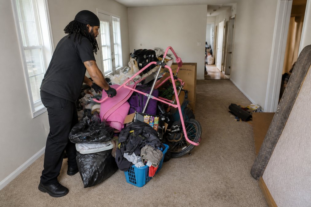 Homeowner clearing a pathway through a cluttered living room with furniture stacked and marked for removal, showing accessibility preparation with open doorways and cleared hallways, natural daylight from windows