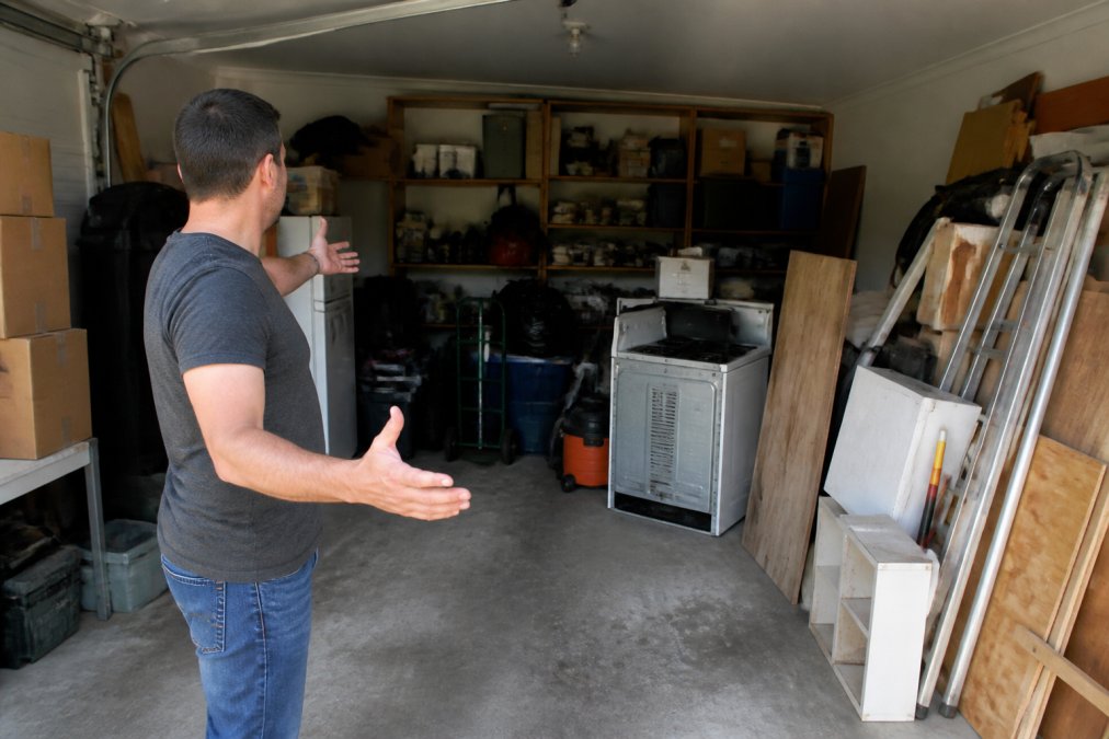 Homeowner standing in cluttered garage filled with stacked boxes, old furniture, appliances, and construction debris, gesturing to show the volume of items needing removal